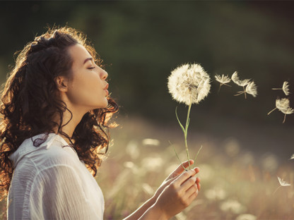 Młoda kobieta na polu dmuchająca dmuchawca, wokół którego unoszą się pyłki, alergen, alergia na pyłki traw