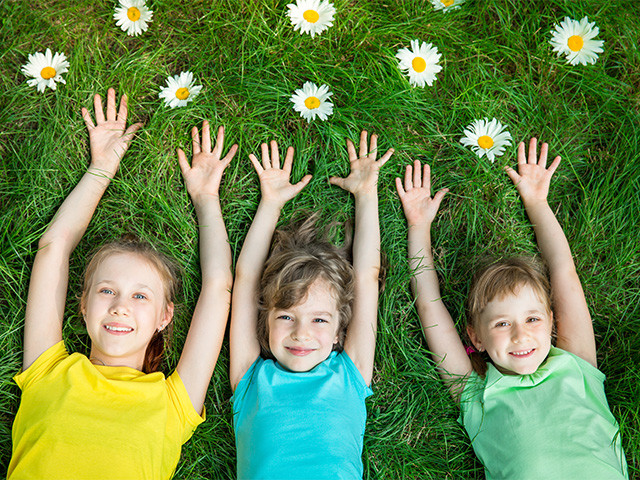 Trzy dziewczynki leżą na trawie wśród stokrotek z uniesionymi rękami i uśmiechami